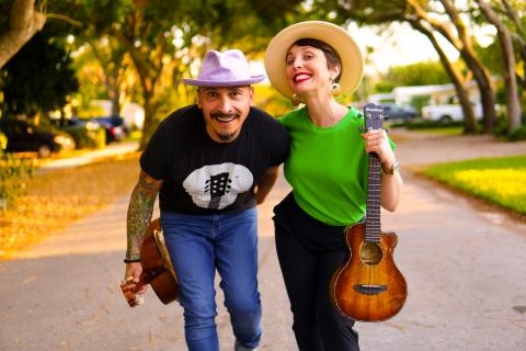 A A man and a woman walking while holding a guitar and wearing hats.