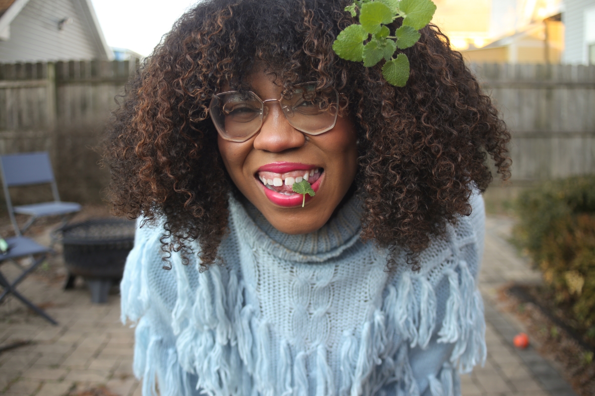Smiling black woman with glasses. light blue sweater, and mint leaves in her mouth and hair